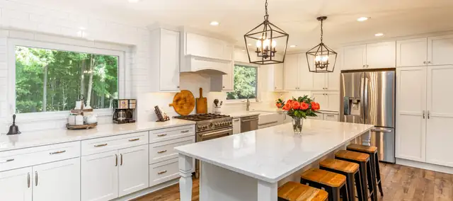 White kitchen by MJ Kitchen & Bath with a marble-look island, wood counter stools and three lantern pendants