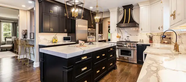 Kitchen by MJ Kitchen & Bath with black cabinets, a white marble-look island, brass pendant lights and a black range hood with brass trim