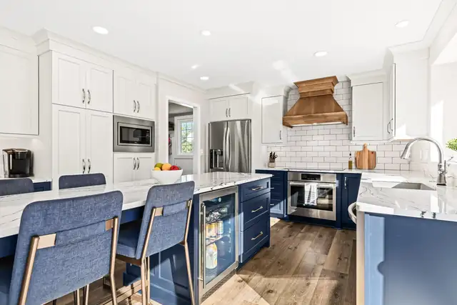 White kitchen with a navy blue island, blue upholstered counter stools and a copper range hood